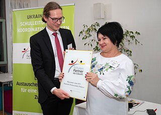 Übergabe der PASCH-Plakette Ein Mann im schwarzen Anzug und eine Frau in einem weißen Oberteil stehen vor einem grünen Goethe-Institut-Aufsteller und halten gemeinsam eine Plakette, die die Aufschrift "Schulen: Partner der Zukunft" trägt.